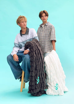 Two models pose with two thick, Minky Couture Dude Perfect Hugs blankets in a studio setting. The model on the left sits on a wooden stool wearing a light blue and gray color-blocked long-sleeve shirt and blue jeans, while the model on the right stands in a brown and beige plaid short-sleeve button-up shirt.