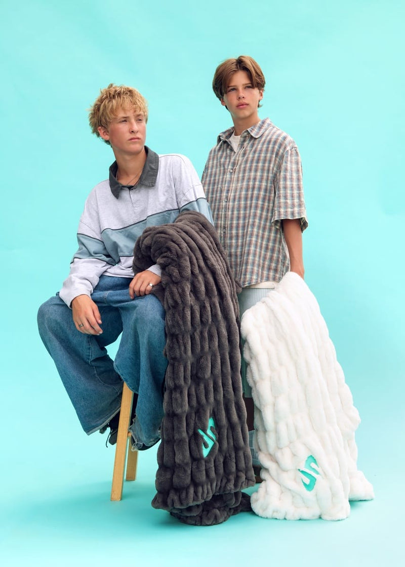 Two models pose with two thick, Minky Couture Dude Perfect Hugs blankets in a studio setting. The model on the left sits on a wooden stool wearing a light blue and gray color-blocked long-sleeve shirt and blue jeans, while the model on the right stands in a brown and beige plaid short-sleeve button-up shirt.