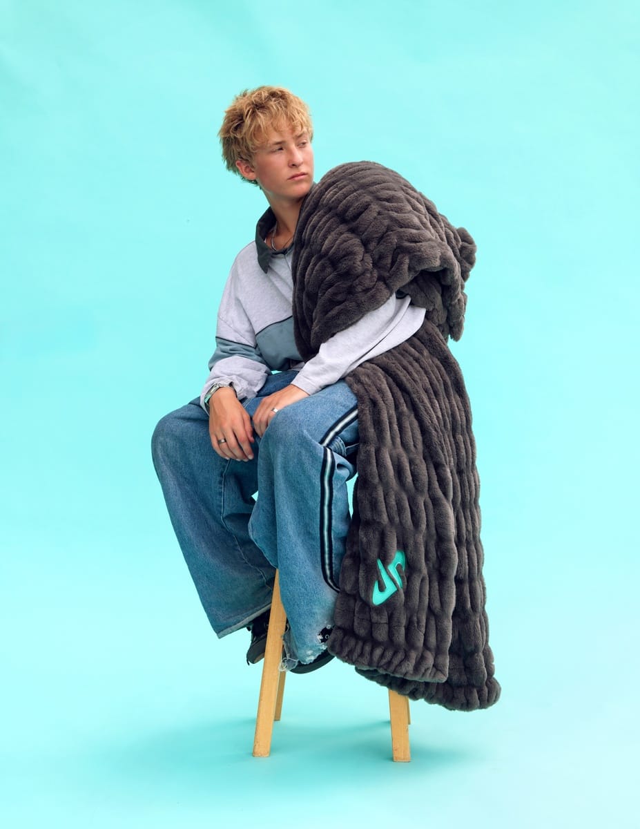  A model sits on a wooden stool against a solid light blue background, wrapped in a thick Minky Couture Dude Perfect Hugs Charcoal blanket. The model is wearing a gray and blue color-blocked long-sleeve shirt and blue jeans.