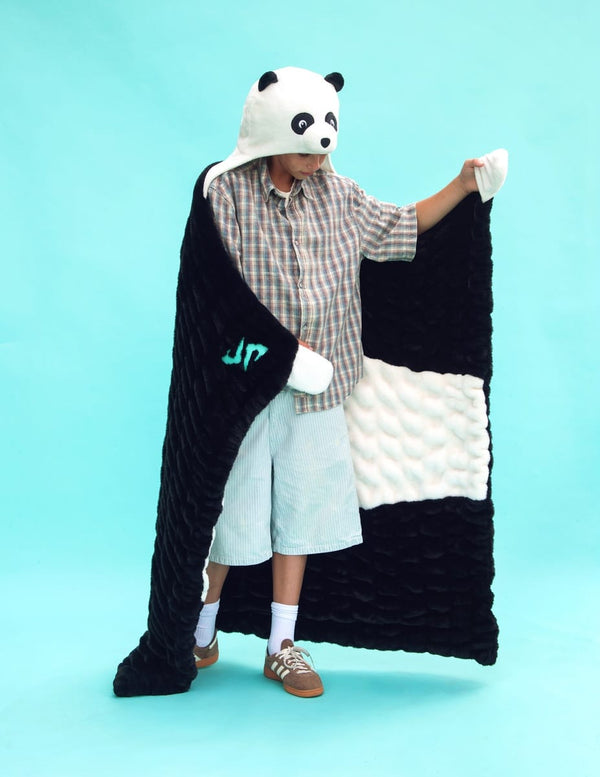 A child modeling a plush black and white panda-themed Dude Perfect blanket with a hood shaped like a panda face, standing against a solid light blue studio background.
