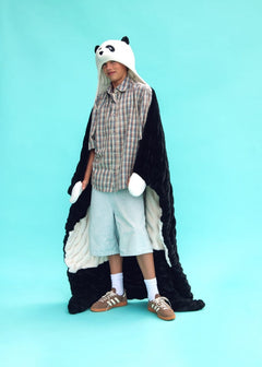 A person wearing a plush panda hooded huggable Dude Perfect blanket with white gloves, a plaid shirt, light blue shorts, white socks, and brown sneakers, standing against a solid light blue background.