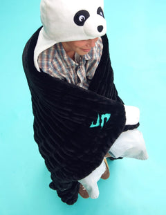 A child wrapped in a black plush panda-themed Dude Perfect blanket, wearing a white hooded panda hat and a plaid shirt, set against a solid light blue background.