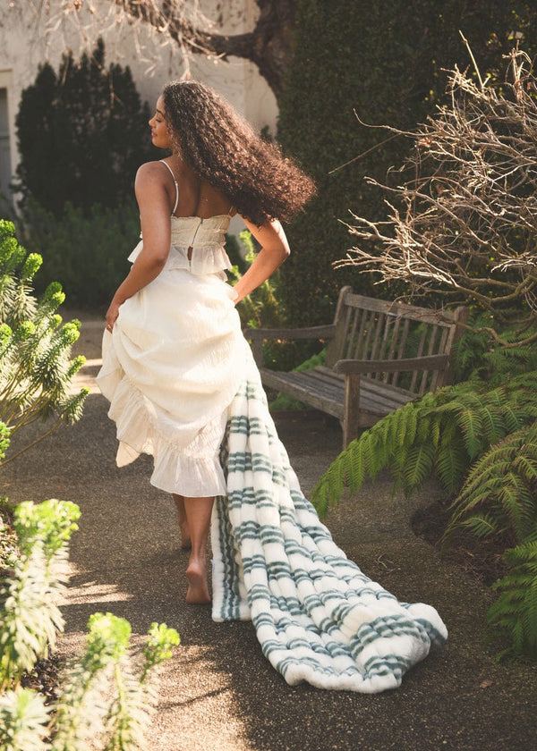 A model wearing a white dress walks away, showcasing the Hugs French Stripe Blanket with blue stripes in a garden setting.