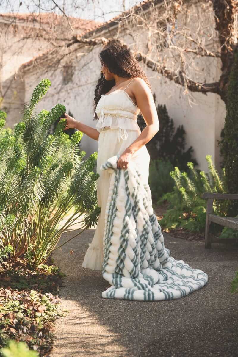 Model draped in the Hugs French Stripe Blanket in a garden, showcasing its plush and cozy design.