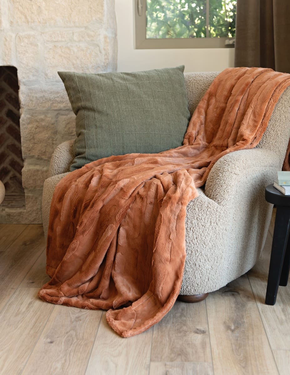 A rust-orange plush Minky Couture Adobe Sorbet throw blanket is draped over a cream boucle armchair in a cozy living room setting with a green pillow, stone wall, and wood floor.