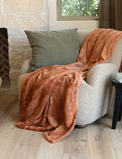 A rust-orange plush Minky Couture Adobe Sorbet throw blanket is draped over a cream boucle armchair in a cozy living room setting with a green pillow, stone wall, and wood floor.