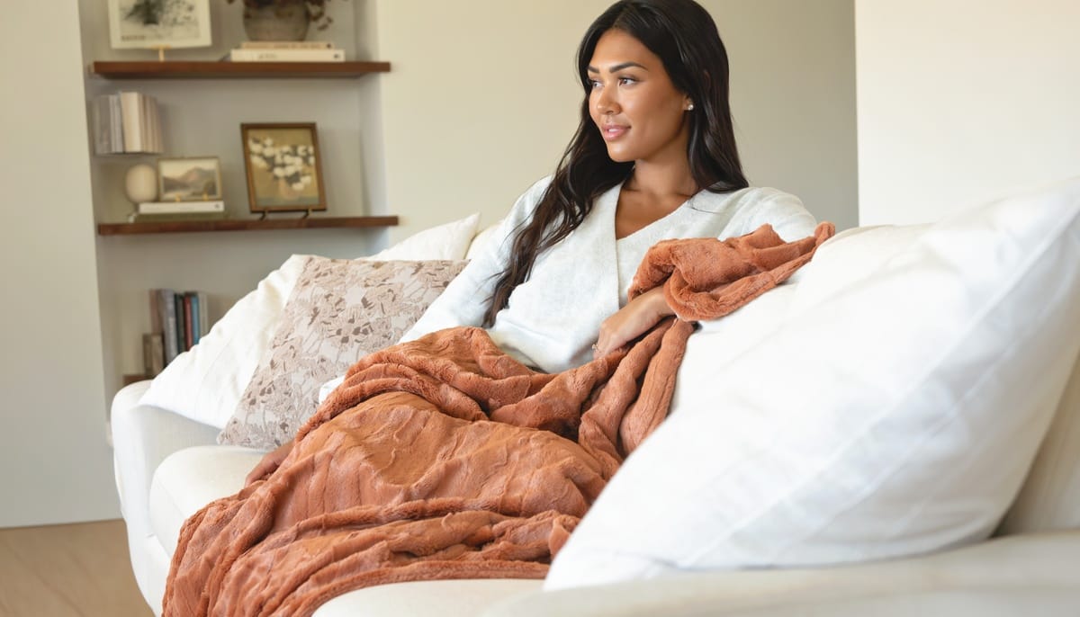 A woman sits on a white sofa in a living room, wrapped in a rust-colored Minky Couture Adobe Sorbet textured throw blanket.