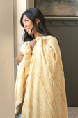 A woman with long dark hair is shown from the side, wrapped in a soft, textured Sorbet Butter Yellow blanket. She is standing indoors next to a light-colored wall and a dark wood cabinet.