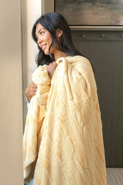 A woman with long dark hair is shown from the side, wrapped in a soft, textured Sorbet Butter Yellow blanket. She is standing indoors next to a light-colored wall and a dark wood cabinet.