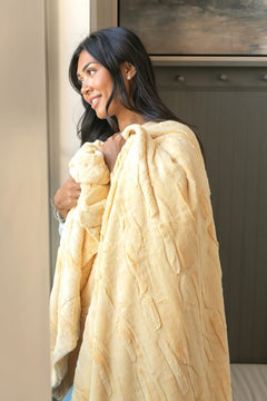 A woman with long dark hair is shown from the side, wrapped in a soft, textured Sorbet Butter Yellow blanket. She is standing indoors next to a light-colored wall and a dark wood cabinet.