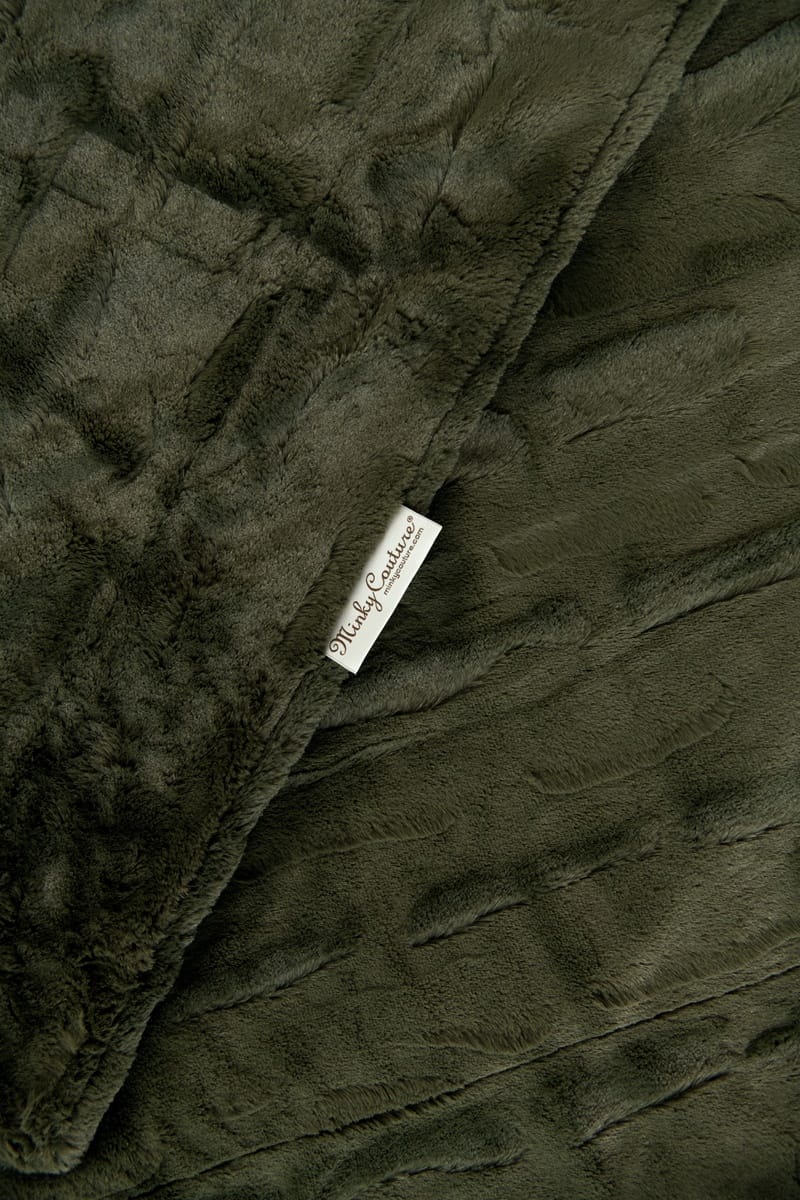 A close-up of a Minky Couture Sorbet Deep Moss  blanket with a textured quilted pattern and a small white brand tag reading 'Mildly Comfortable'.