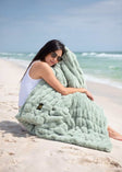 A woman relaxing on the beach with a Hugs Mist Blanket in soothing sage green.