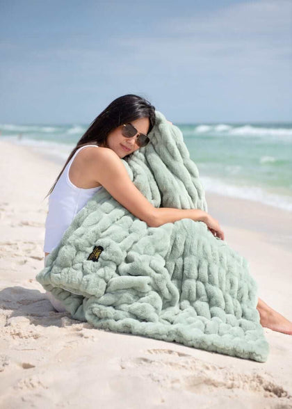 A woman relaxing on the beach with a Hugs Mist Blanket in soothing sage green.