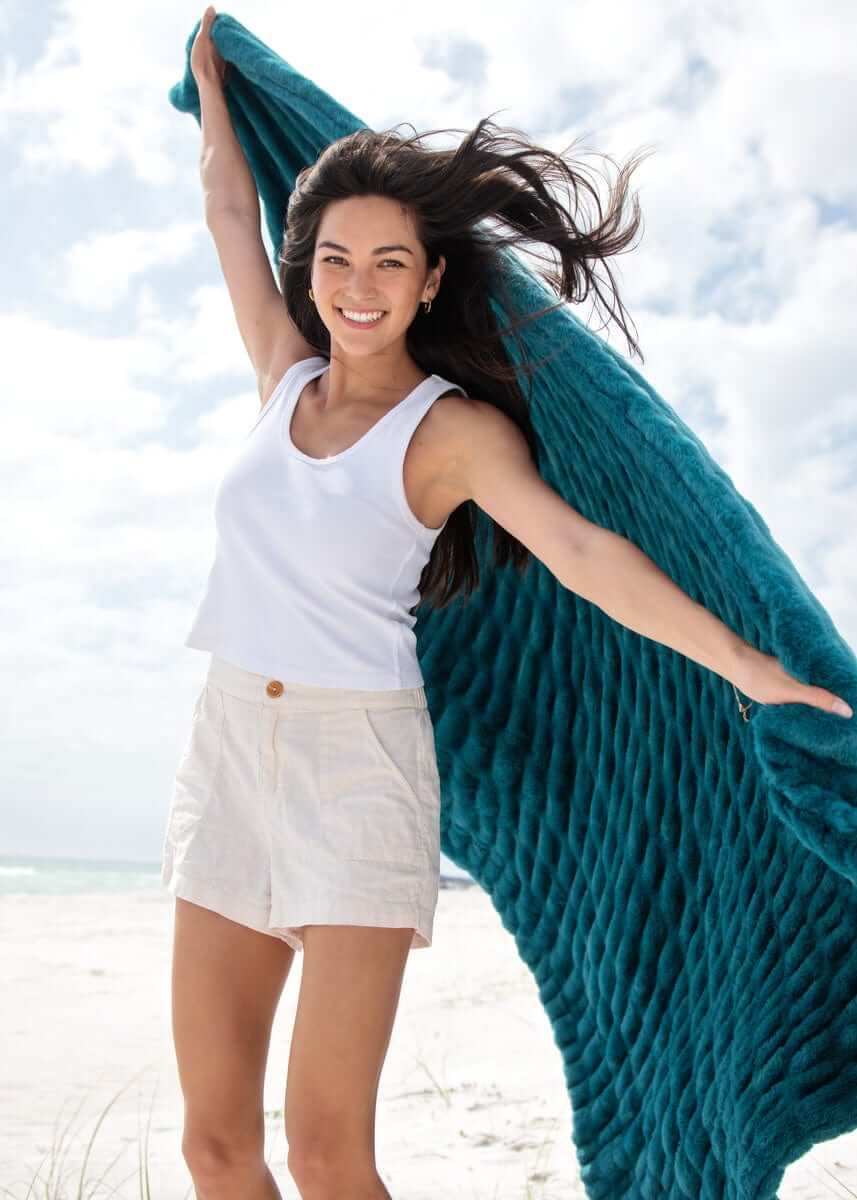 Woman joyfully displaying the Hugs Deep Lake Blanket on a beach with a vibrant turquoise color and sunny sky.