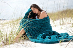 A woman wrapped in the Minky Couture Hugs Deep Lake Blanket, enjoying comfort on a sandy beach.