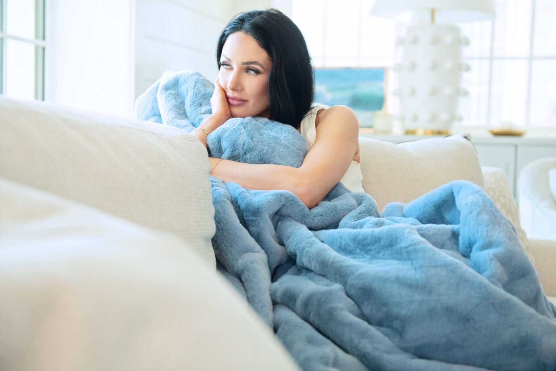 A woman cozying up with a Lush Denim Blanket on a couch, showcasing its plush texture and denim blue color.