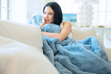 A woman cozying up with a Lush Denim Blanket on a couch, showcasing its plush texture and denim blue color.