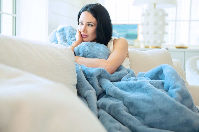 A woman cozying up with a Lush Denim Blanket on a couch, showcasing its plush texture and denim blue color.