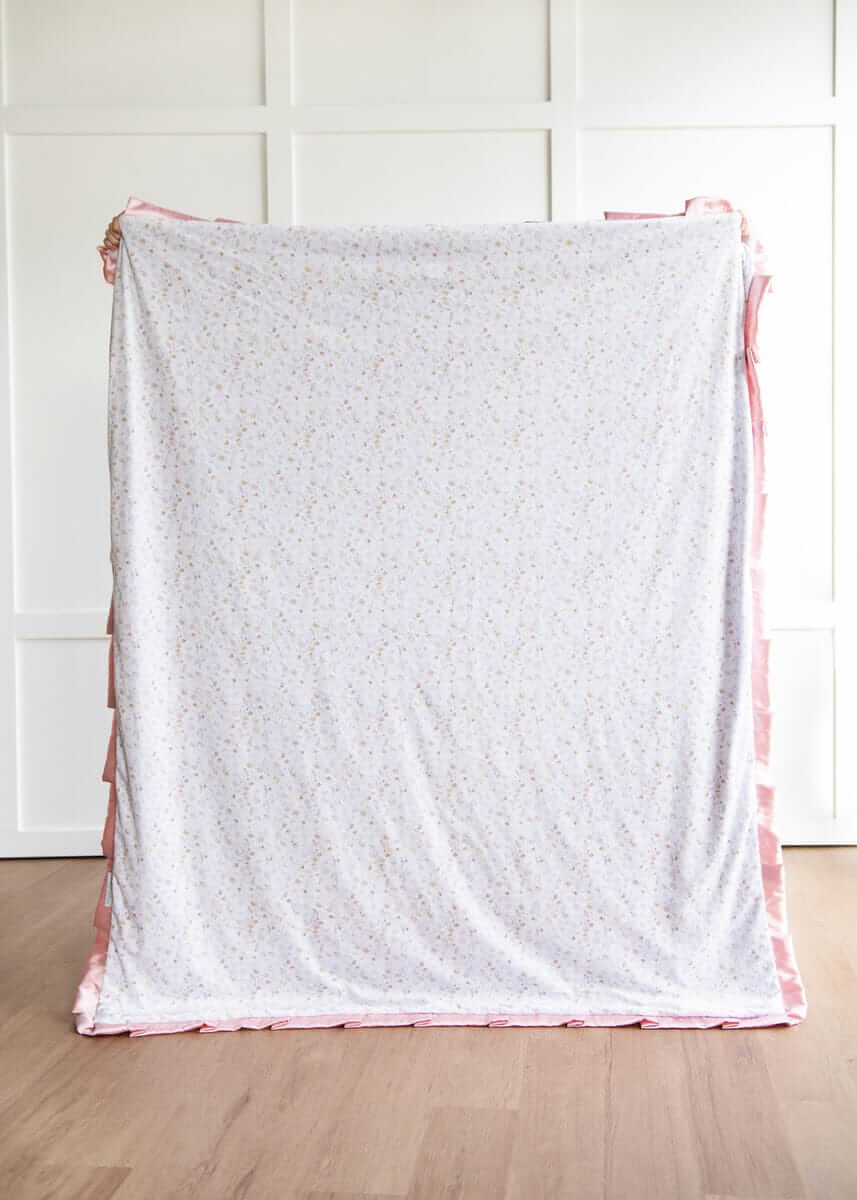 French floral patterned blanket with pink trim on a wooden floor against a white paneled wall.