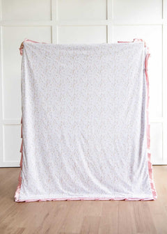 French floral patterned blanket with pink trim on a wooden floor against a white paneled wall.