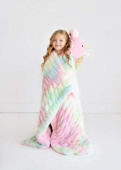 A girl wrapped in the Huggable Unicorn blanket, holding the unicorn head hood, showcasing its pastel tie-dye design.