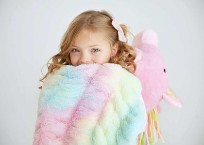 A child wrapped in the Huggable Unicorn pastel tie-dye blanket with a plush unicorn toy.