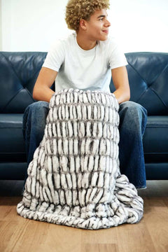 Person sitting on a couch wrapped in the Hugs Obsidian Blanket made of soft faux fur.