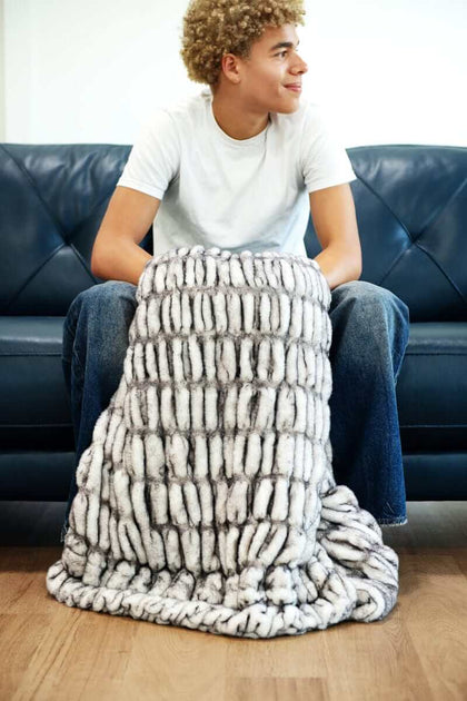 Person sitting on a couch wrapped in the Hugs Obsidian Blanket made of soft faux fur.