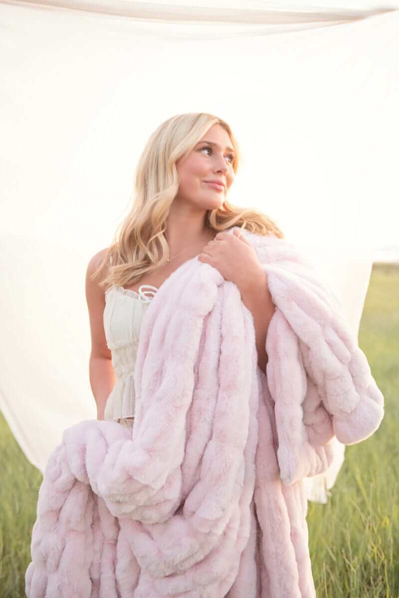 Woman holding the Hugs Bashful Pink Blanket in a scenic field, showcasing its plush texture and soft color.