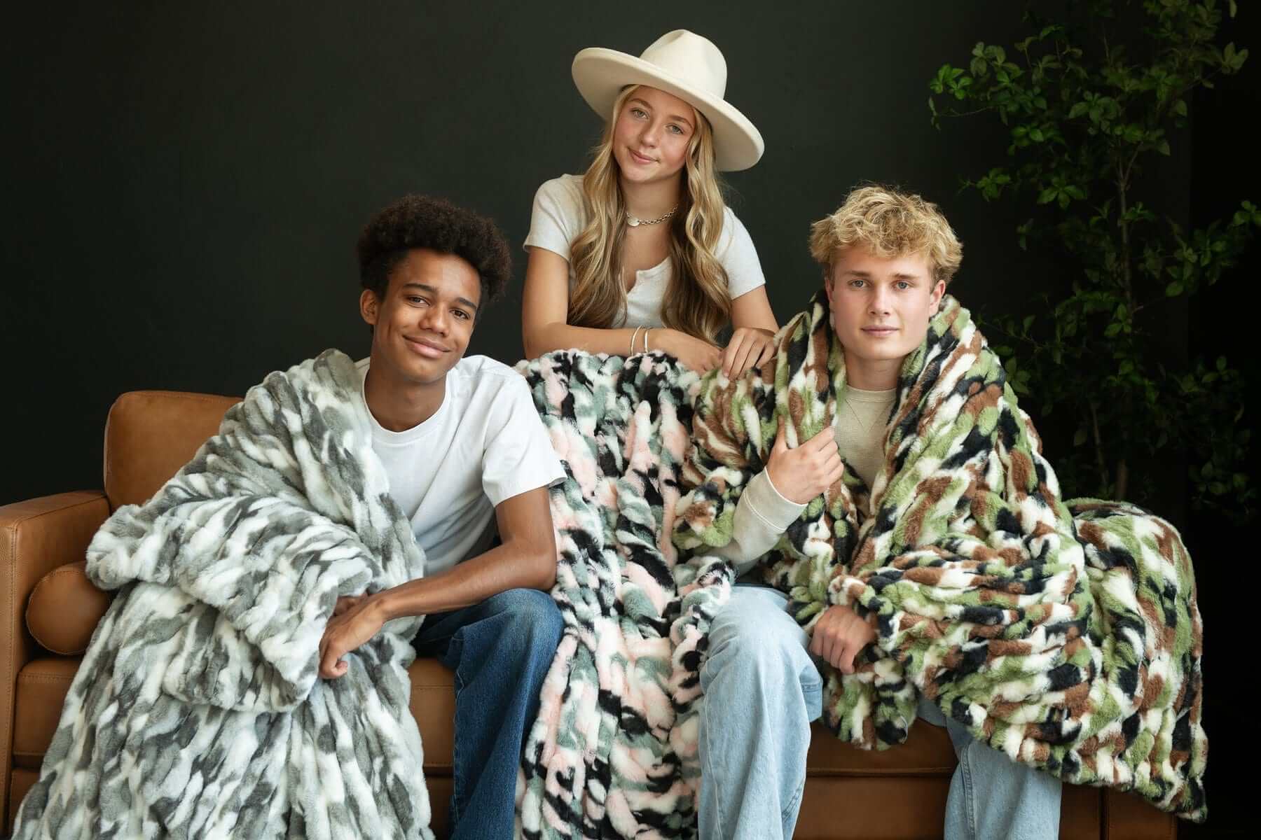 Three people wrapped in stylish blankets, showcasing the Hugs Camo Green Blanket for comfort and chic appeal.