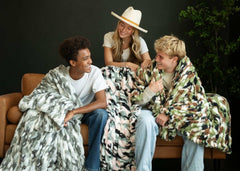 Three friends enjoying time together, draped in cozy blankets, featuring the Hugs Camo Green Blanket and playful patterns.