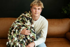 Hugs Camo Green Blanket wrapped around a young man lounging on a couch, showcasing its stylish camo design.