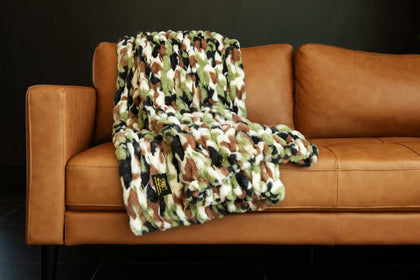 Hugs Camo Green Blanket draped over a stylish leather couch, showcasing its vibrant camo pattern.