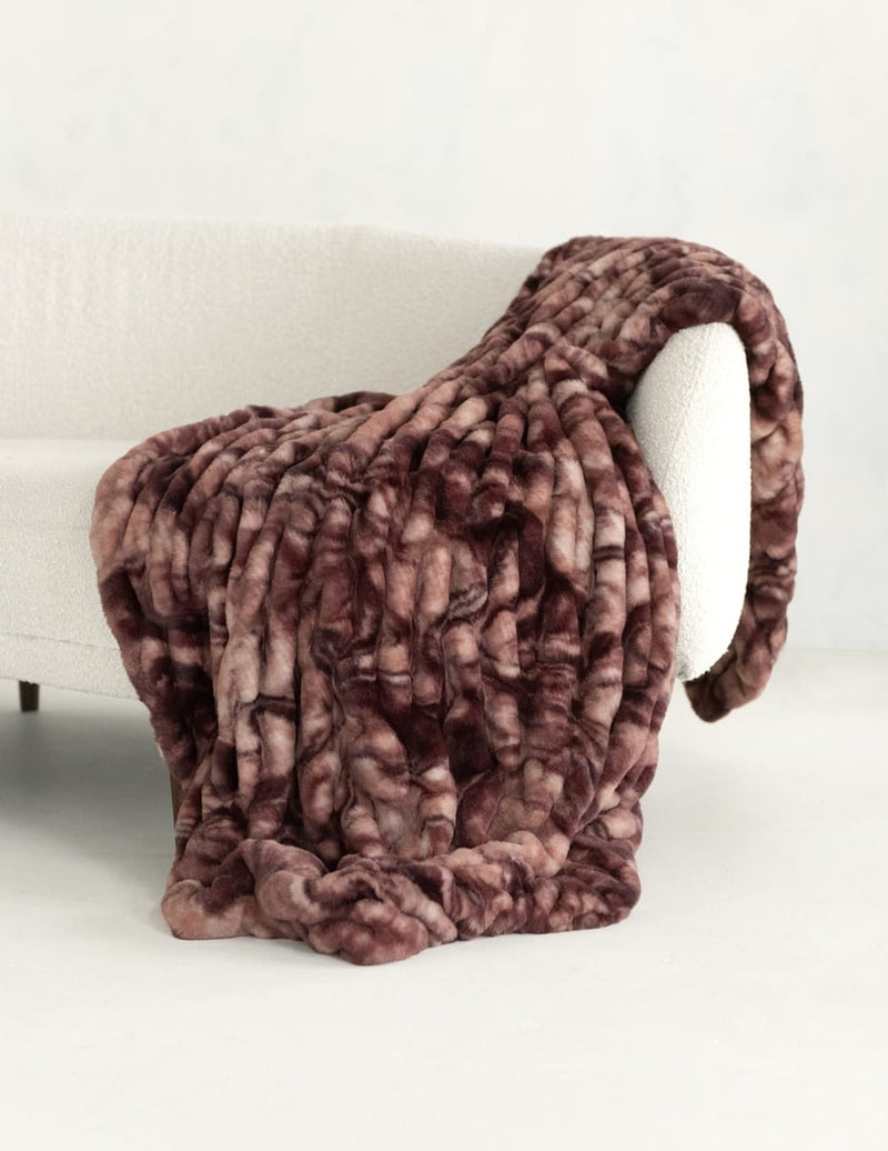 A chunky, plush faux fur throw blanket with a mottled brown and dusty rose pattern, draped over a white armchair in a minimalist room setting.