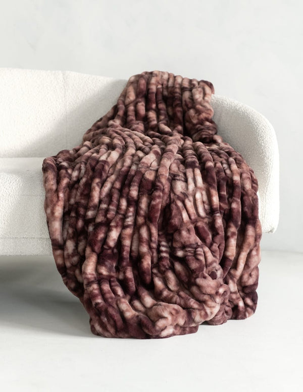 A chunky throw blanket with a marbled pattern in dusty rose, mauve, and brown, draped over a white textured armchair.