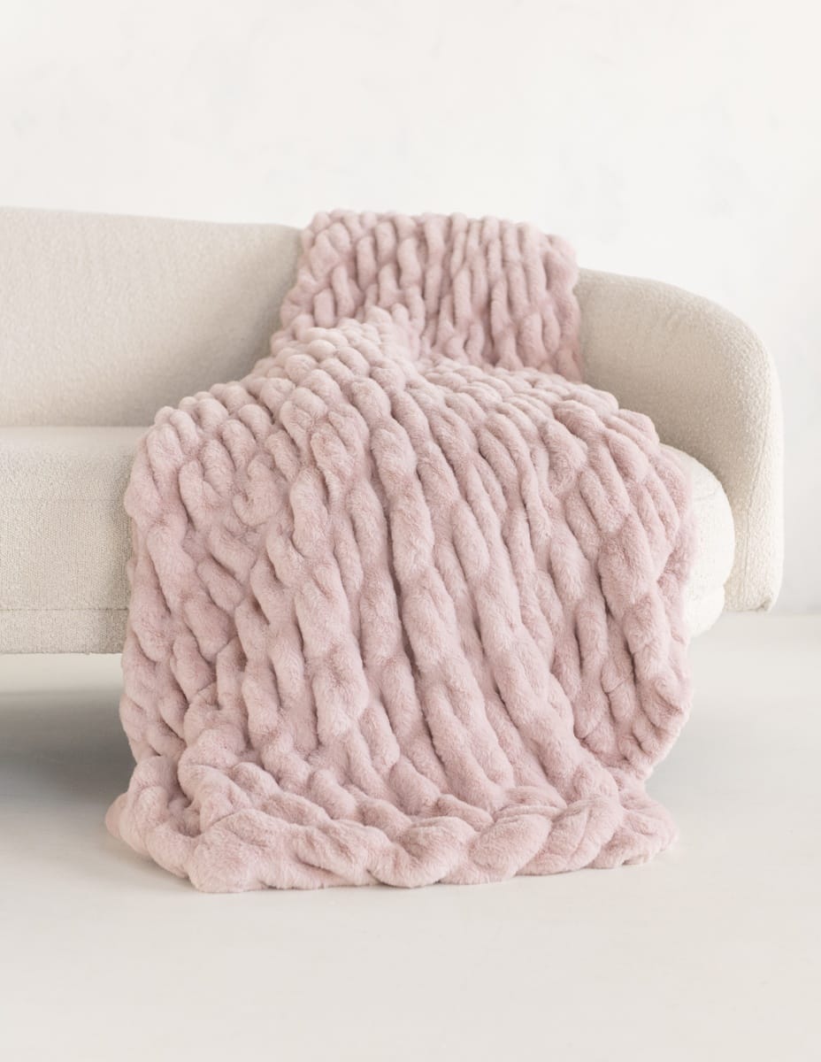 A chunky plush blush pink throw blanket draped over a light beige sofa against a clean white background.