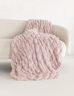 A chunky plush blush pink throw blanket draped over a light beige sofa against a clean white background.
