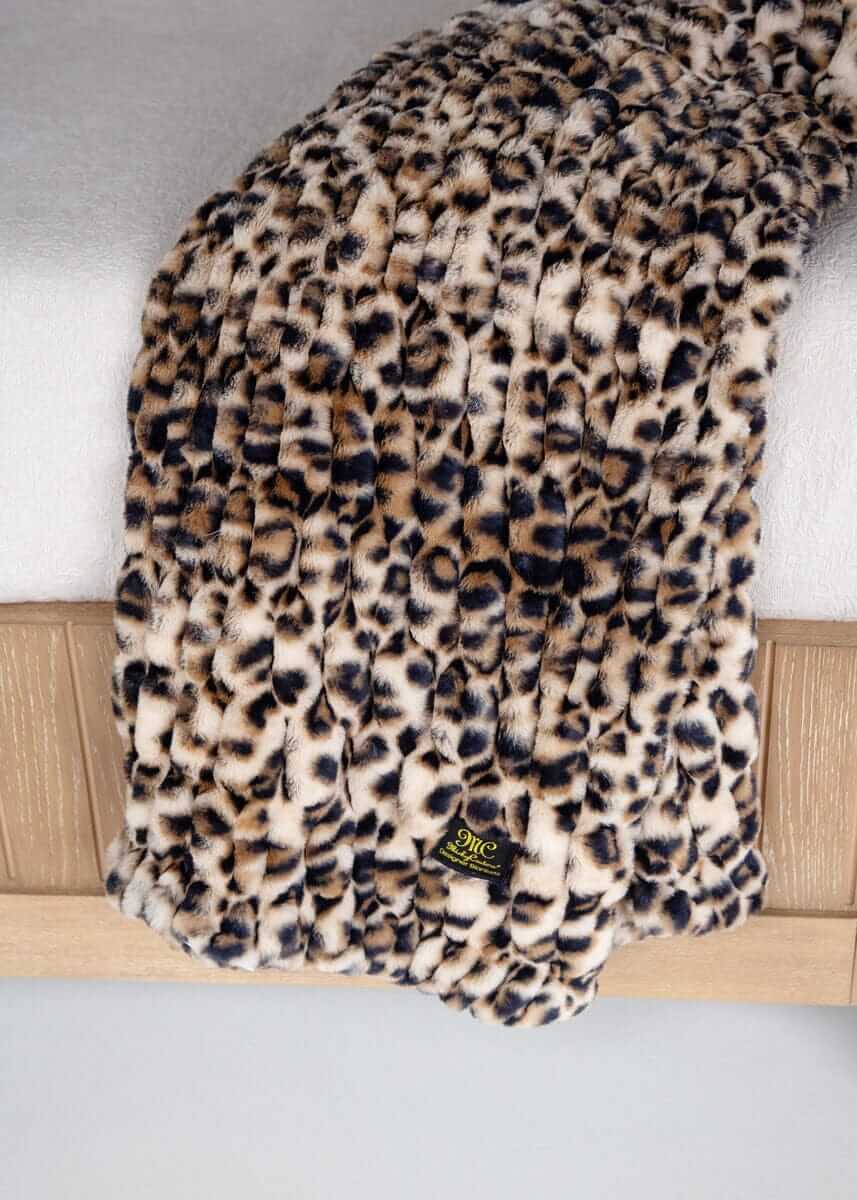 Leopard print Minky Couture faux fur throw blanket draped over a bed with brand tag visible.