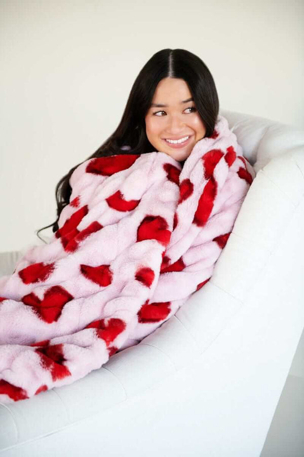 A woman happily snuggled in the Hugs Heart Red Blanket featuring vibrant red hearts on a soft pink background.
