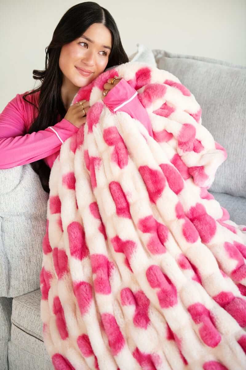 A woman wrapped in the Hugs Heart Pink Blanket on a grey couch, showcasing its plush design and charming pink hearts.