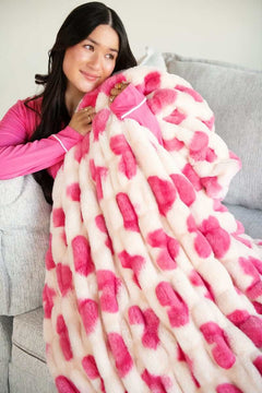 A woman wrapped in the Hugs Heart Pink Blanket on a grey couch, showcasing its plush design and charming pink hearts.