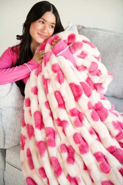 A woman wrapped in the Hugs Heart Pink Blanket on a grey couch, showcasing its plush design and charming pink hearts.