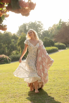 Woman in a light dress joyfully holding the Hugs Polka Dot Blush Blanket outdoors, surrounded by greenery.