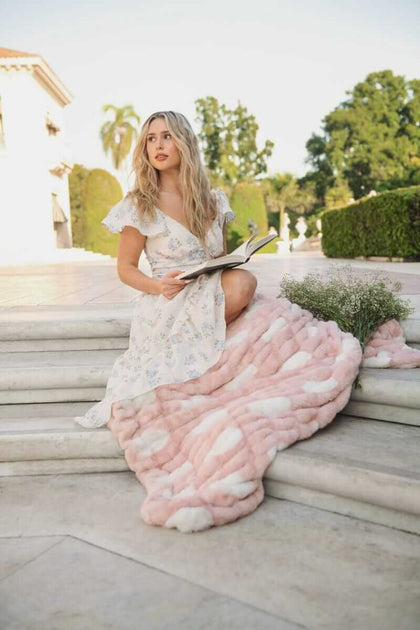 Woman sitting on stairs with Minky Couture Hugs Polka Dot Blush Blanket, reading a book, surrounded by greenery and soft sunlight.