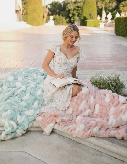 A woman reading a book while seated on the Minky Couture Hugs Polka Dot Tiffany Blanket in Tiffany blue and a pink blanket.