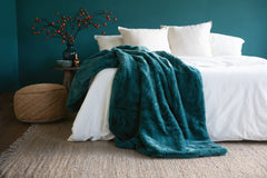 A plush teal throw blanket draped over a neatly made bed with white bedding, set in a bedroom with a dark teal wall, a woven pouf, and a small side table with decorative branches.