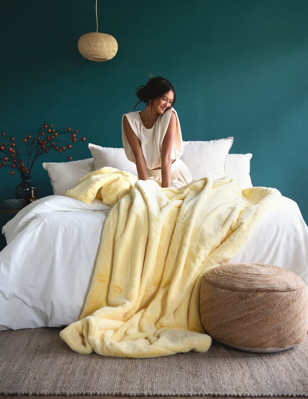 A woman sits on a bed in a styled bedroom, wrapped in a pale yellow plush throw blanket. The bed is dressed in white bedding, set against a deep teal wall.