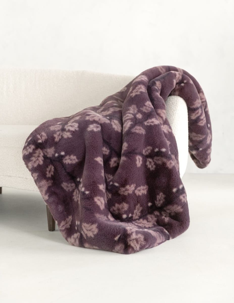 A plush throw blanket in a dusty purple color with a lighter floral pattern, draped over the arm of a light-colored armchair.