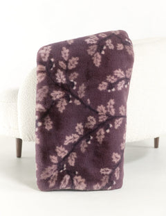 A plush throw blanket with a dark purple background and a light pink leafy branch pattern, draped over the arm of a white textured armchair.