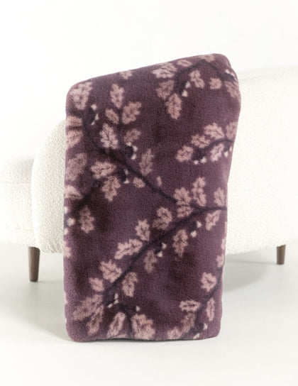 A plush throw blanket with a dark purple background and a light pink leafy branch pattern, draped over the arm of a white textured armchair.